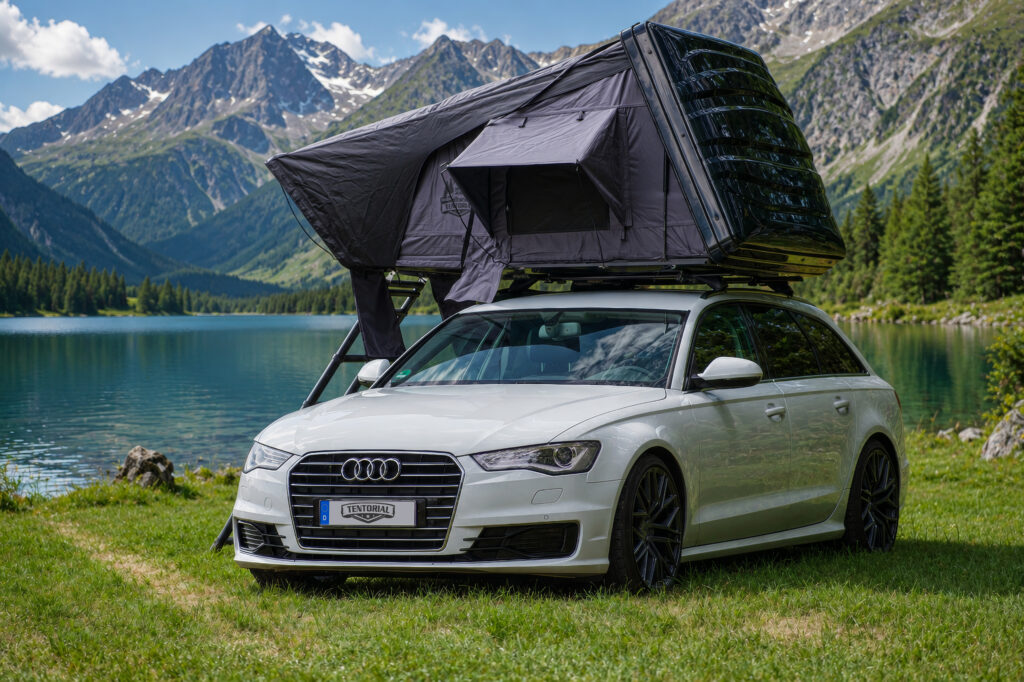 Ein weißer Audi Kombi steht am Bergsee vor den Alpen mit einem schwarzen Hardshell-Dachzelt auf dem Dach.